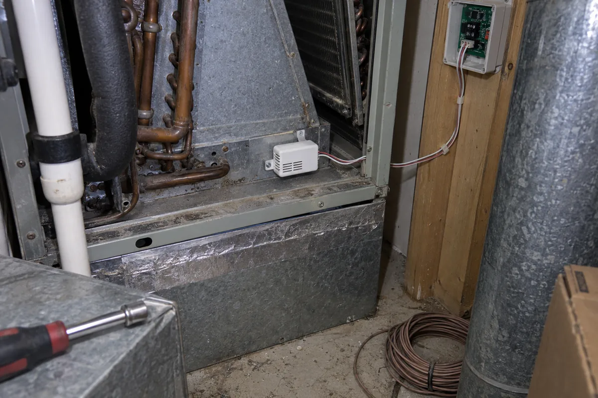 An A2L refrigerant detection sensor mounted low inside an open evaporator coil cabinet in a furnace closet, a two wire harness running from it out to a small control board in a plastic enclosure screwed to a wall stud, with coiled thermostat wire and a nut driver left on the floor.