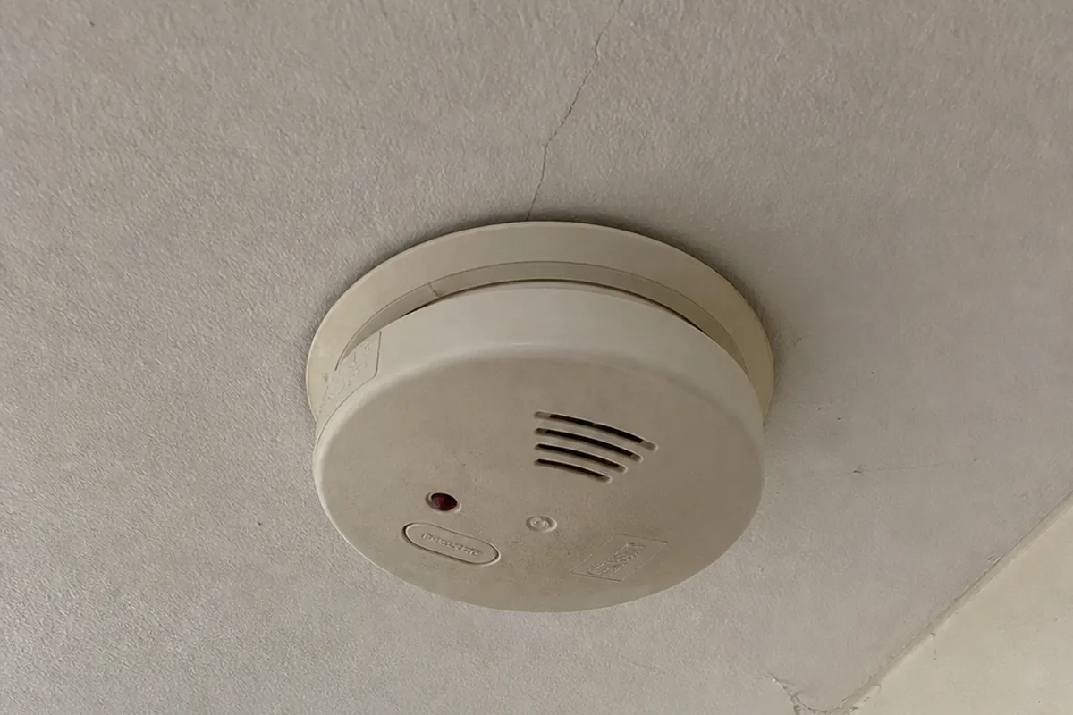 A close view of a round white carbon monoxide alarm mounted on a textured ceiling near a doorway, its red indicator light lit and a hairline crack in the ceiling nearby.