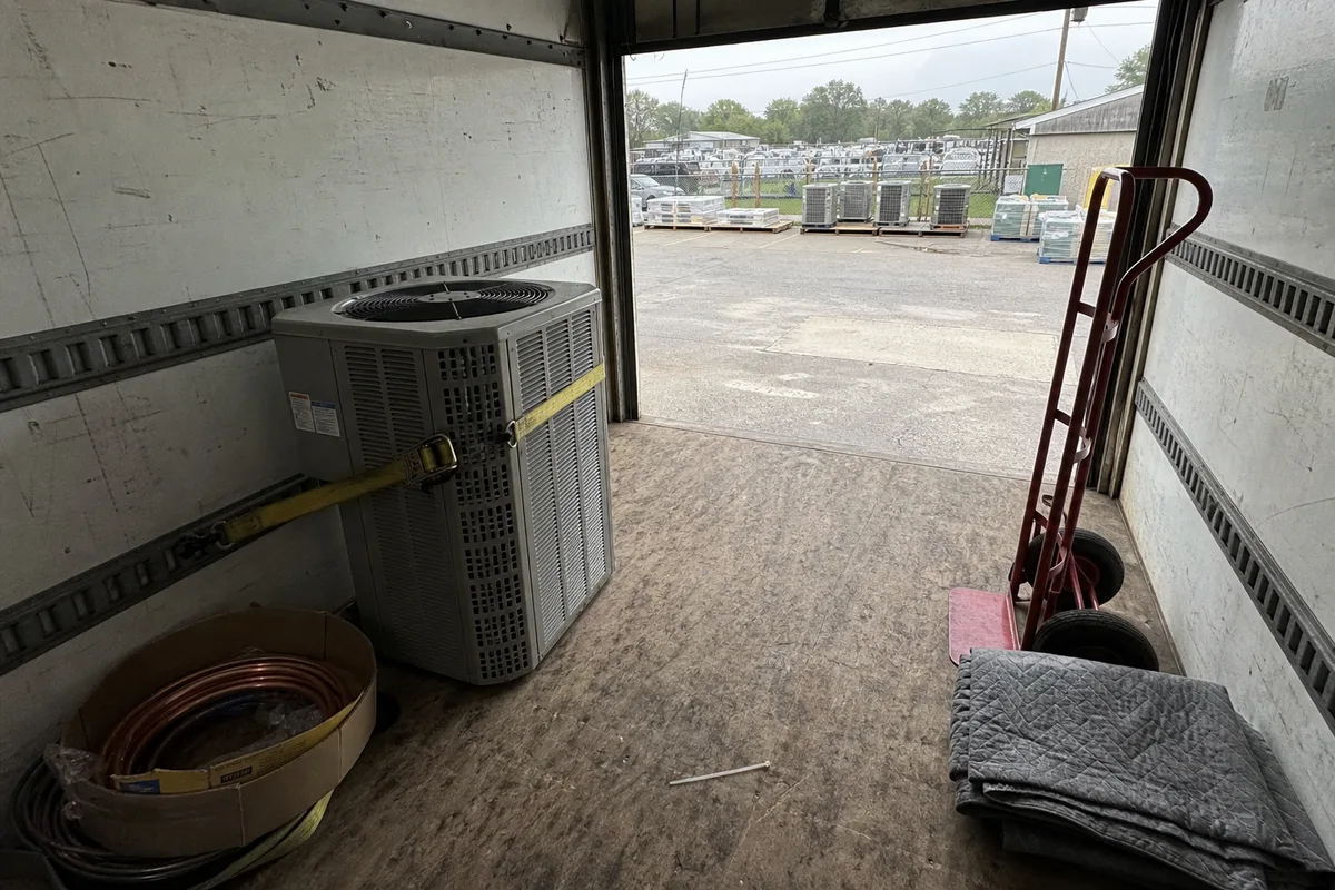 A residential condensing unit strapped upright against the wall inside a box truck, a carton of coiled copper line set on the floor beside it and a red hand truck by the open rear door, with a supply house yard full of palletized condensers on pallets visible outside.