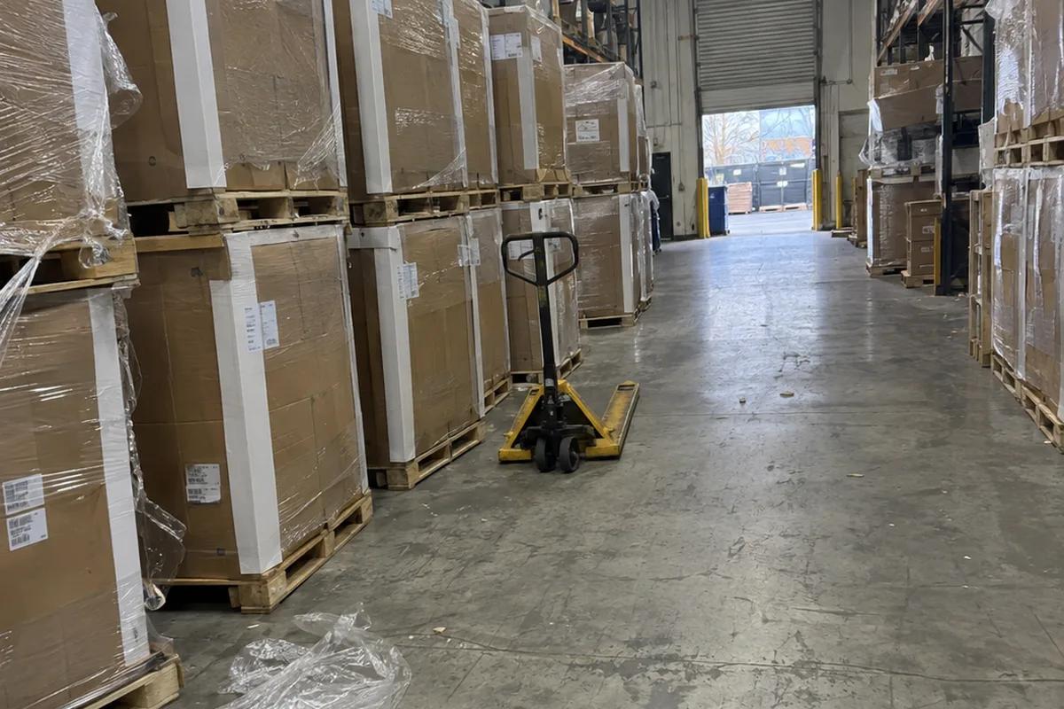 Boxed outdoor condensers stacked on pallets in a distributor warehouse aisle, with a yellow pallet jack, torn shrink wrap on the concrete floor, and a roll-up dock door open at the far end.