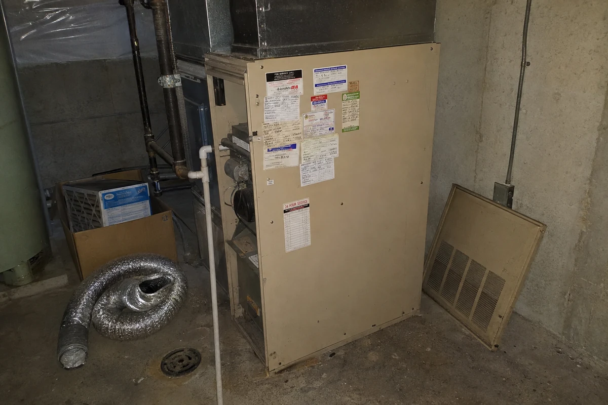 An older gas furnace in a concrete basement, its cabinet covered with layered service stickers and handwritten date tags from past visits, the access panel leaning against the wall beside a coil of flex duct and a box of filters.