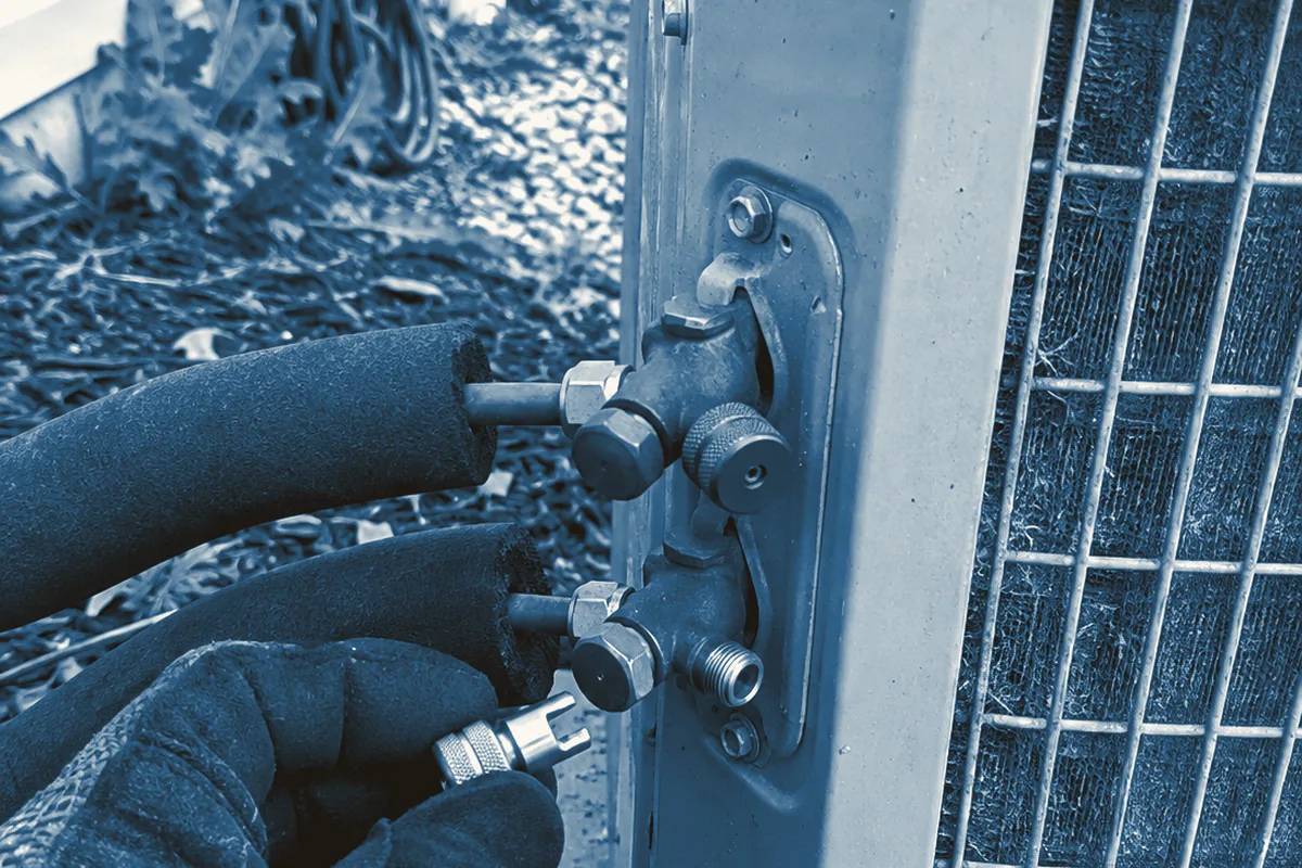 Close view of the brass service valves on the side of a residential condenser, one port fitted with a knurled locking refrigerant cap and the other port bare, a gloved hand holding the cap key beside black foam-insulated line set, mulch and garden hose in the background under flat daylight.