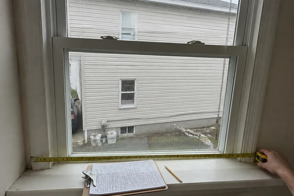 A tape measure held against a window frame inside a house, a clipboard with a handwritten worksheet resting on the sill.